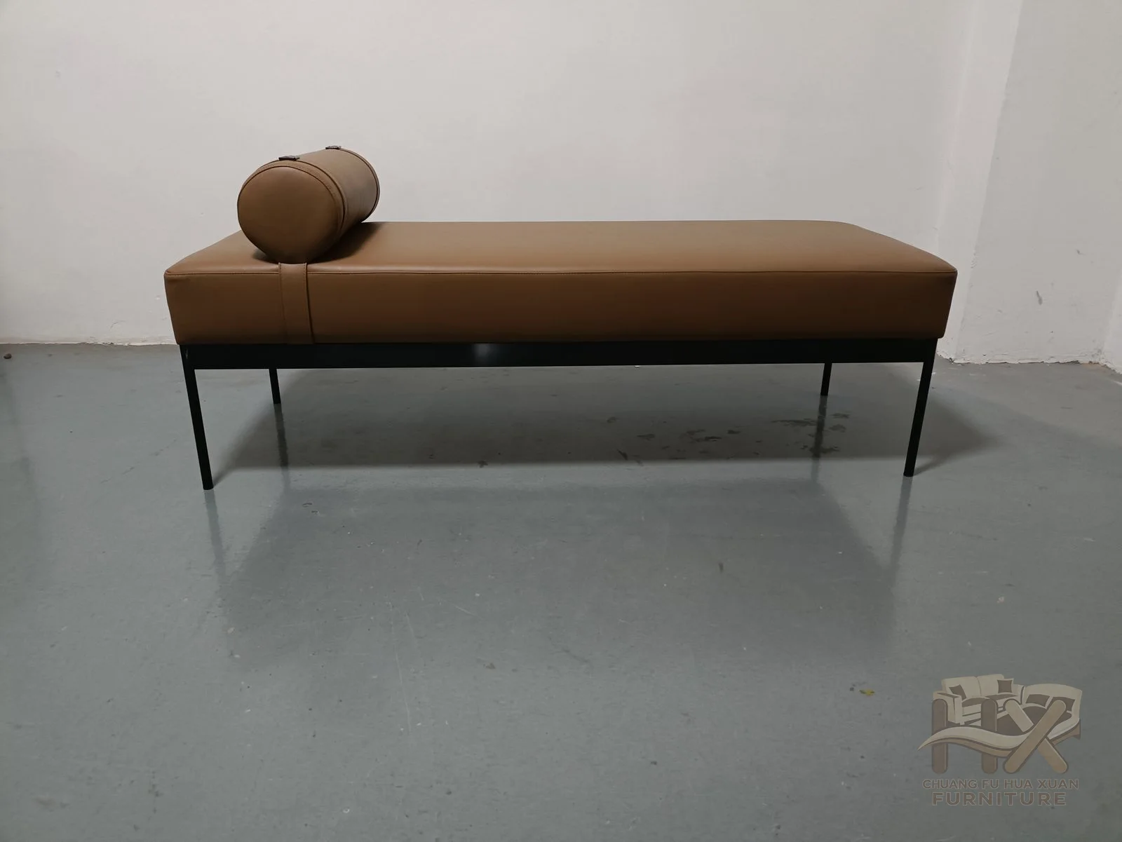 A long tan seat on slender legs carries a low bolster at one end instead of a full upright back.